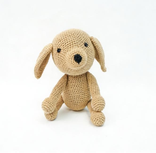 A soft tan crochet puppy sitting patiently on a white background. This floppy-eared amigurumi dog features long ragdoll legs and makes a perfect neutral nursery decor piece.
